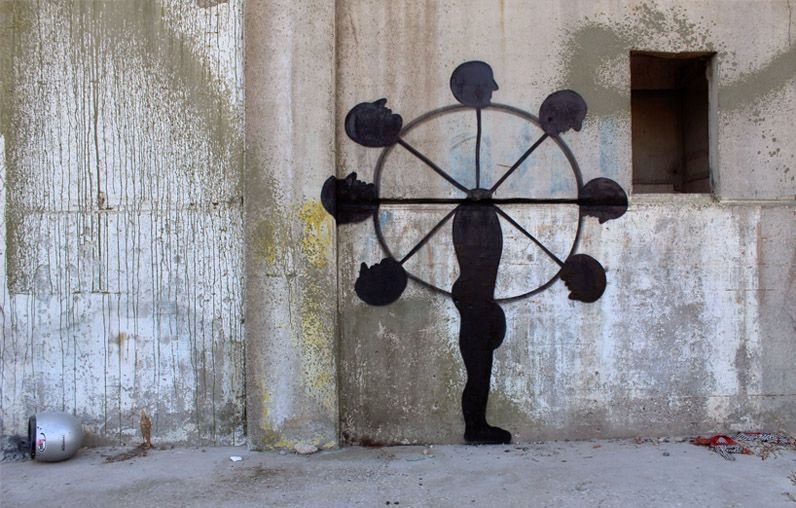 Black silhouette mural of a standing human figure with a wheel of severed heads mounted through its torso like spokes, painted on a weathered concrete wall beside a window opening.