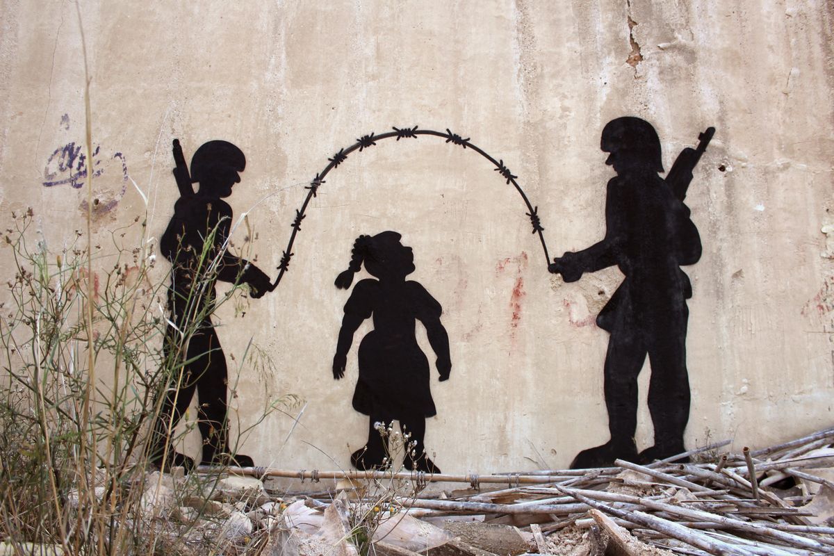 Black silhouette mural of two armed soldiers turning a jump rope made of barbed wire for a small girl skipping between them, painted on a weathered wall surrounded by debris and dry brush.