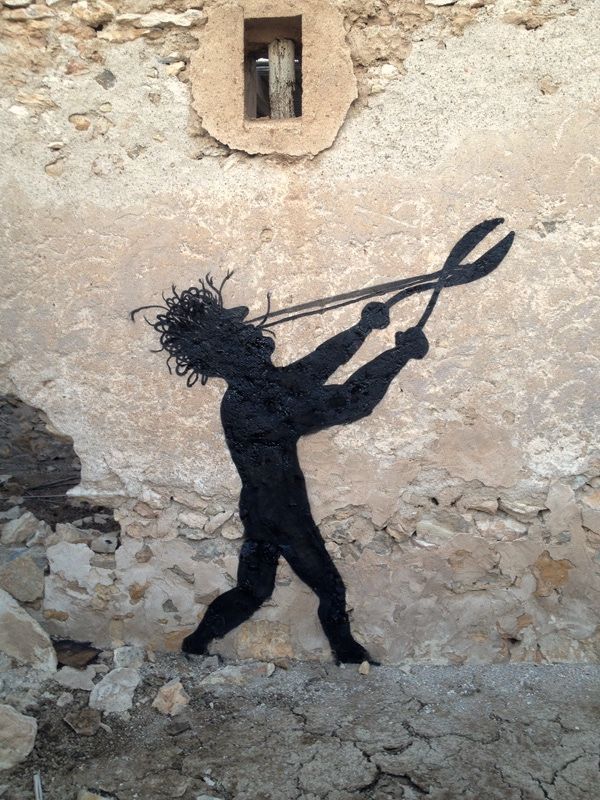 Black silhouette mural of a wild-haired nude figure wielding an oversized pair of garden shears, painted on a rough stone wall beneath a small window opening.
