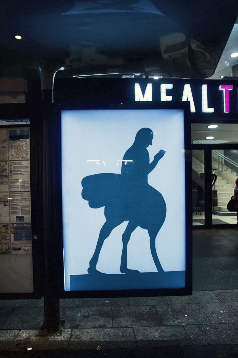 Black silhouette illustration on an illuminated bus shelter ad panel at night, showing a person looking at a phone while riding an ostrich-like bird, a lit shop sign reading 'MEALT' visible behind.