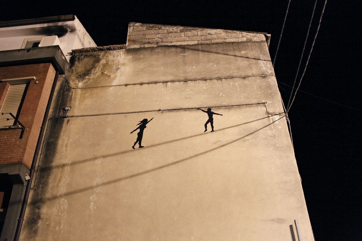 Night view looking up at a building corner mural of two black silhouette tightrope walkers on overhead wires, their long shadows cast across the wall.