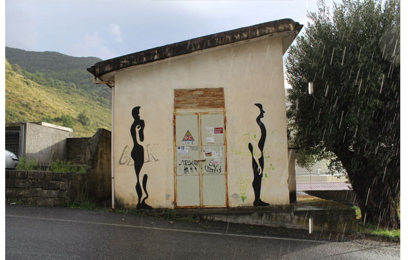 Small rural building with two tall, elongated black silhouette figures painted on either side of a rusted metal door, photographed in the rain with green hills behind.