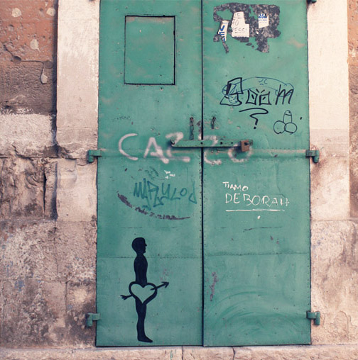 Green metal double door with a black silhouette figure holding a heart between its legs, graffiti tags and handwritten names scrawled across the weathered surface.