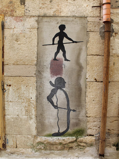 Two black silhouette figures painted in a stone wall niche, one standing above holding a spear, the other crouched below drawing a bow, a rust-stained drainpipe running alongside.