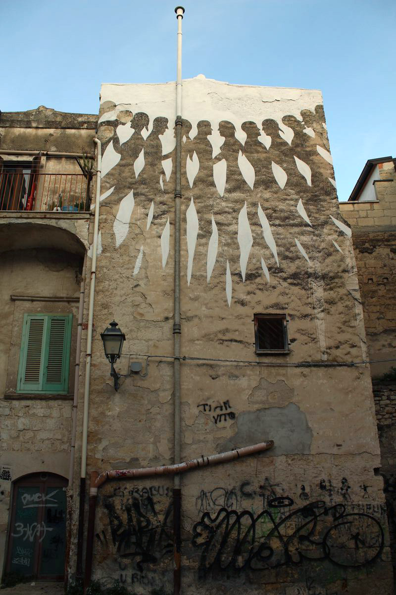 Black-and-white mural on a building facade showing a row of profile figures marching in a chain, each linked by a leaf-shaped white gap, above a window and a drainpipe.