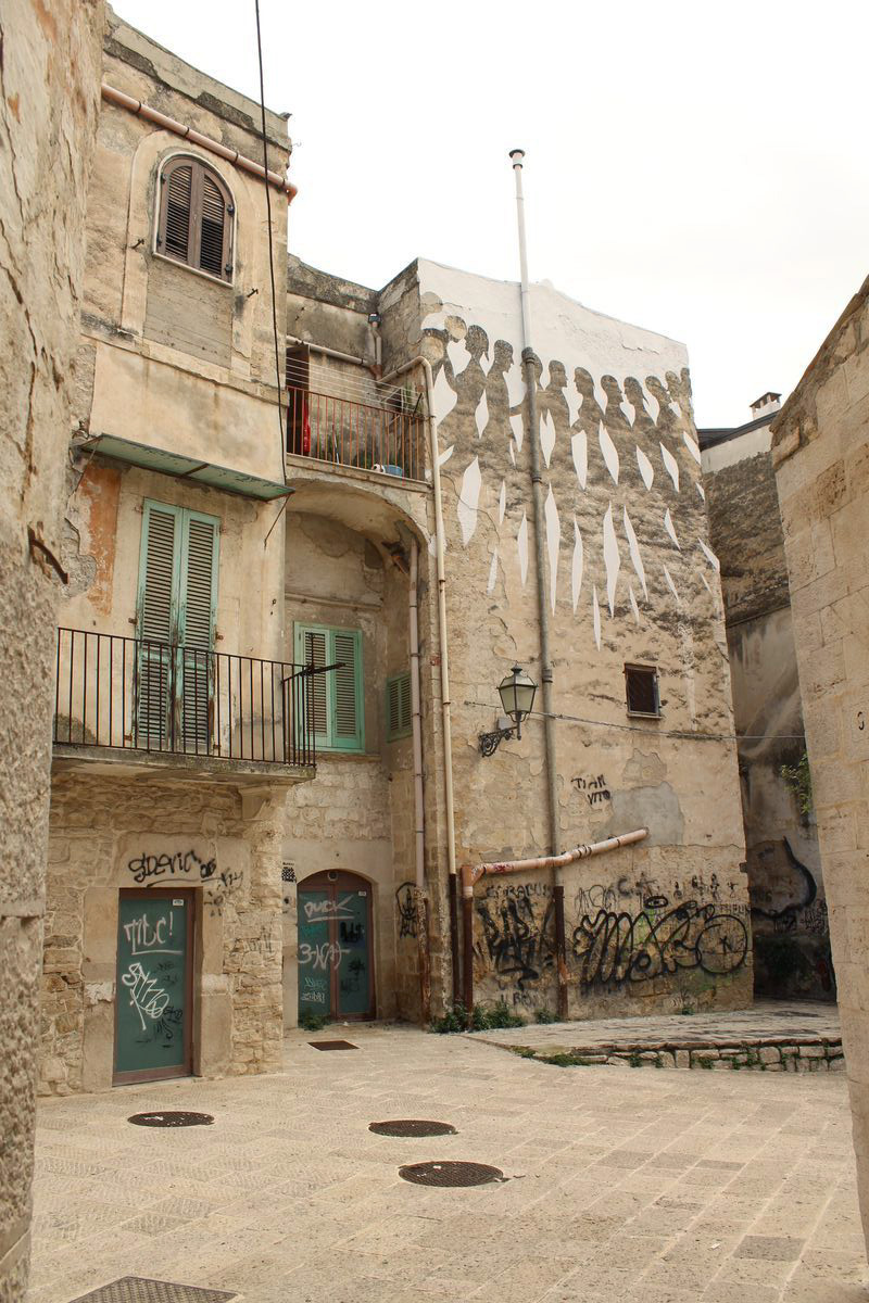 Wide street view of the same building corner mural showing the row of marching profile figures at the top of the facade, with balconies, shuttered windows, and graffiti-covered lower walls, a plaza with manhole covers in the foreground.