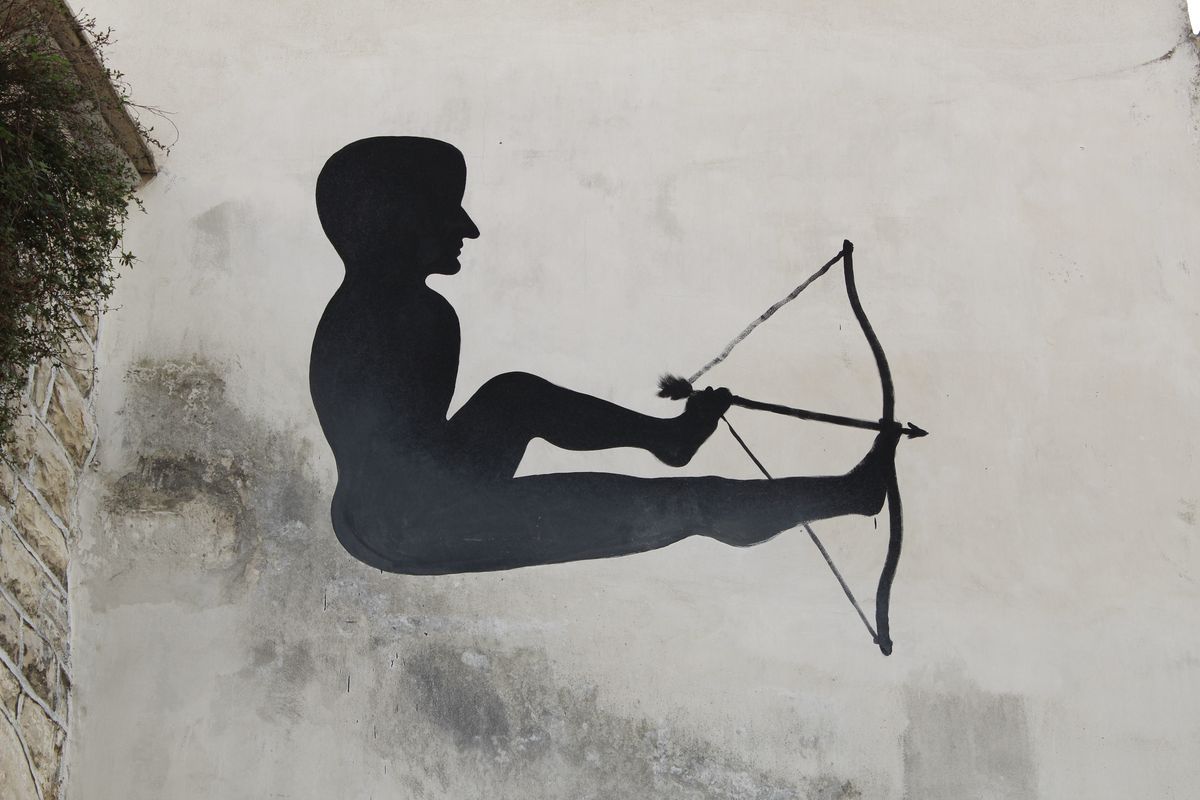 Black silhouette mural of a reclining nude figure with legs raised, using their feet to draw back a large bow and shoot an arrow, painted on a plastered wall beside climbing vines.