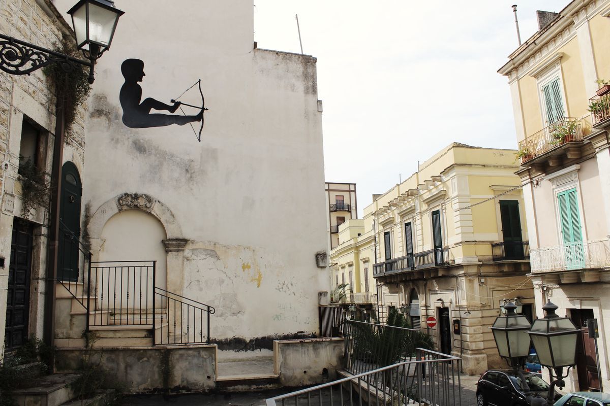 Wide view of the same reclining archer mural high on a building corner, seen from a street lined with lampposts, staircases, and colorful shuttered facades.