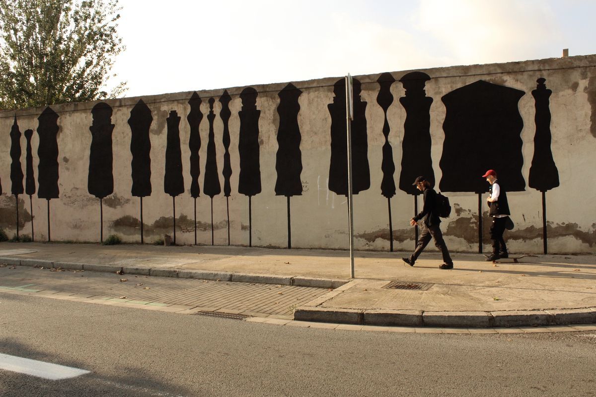 Row of tall black silhouette chess-piece shapes mounted on poles along a plastered wall, two pedestrians walking past on the sidewalk below.