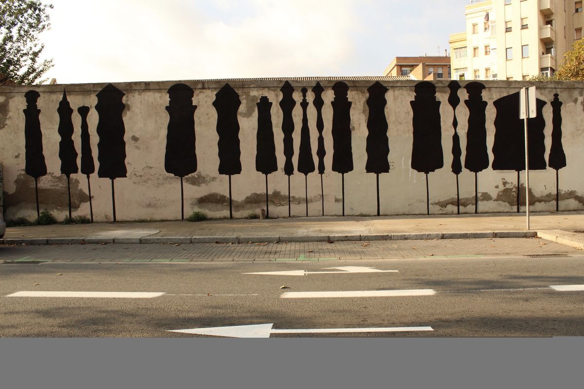 Corner view of four black silhouette chess-piece shapes on poles against a weathered wall, a woman cycling past with a child in a rear seat, parked cars and a no-entry sign nearby.