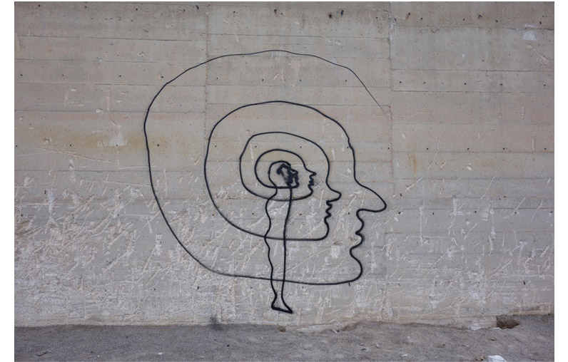 Thin black outline mural on a concrete wall showing a spiral, shell-shaped head containing a smaller profile head and a standing human figure inside the spiral.