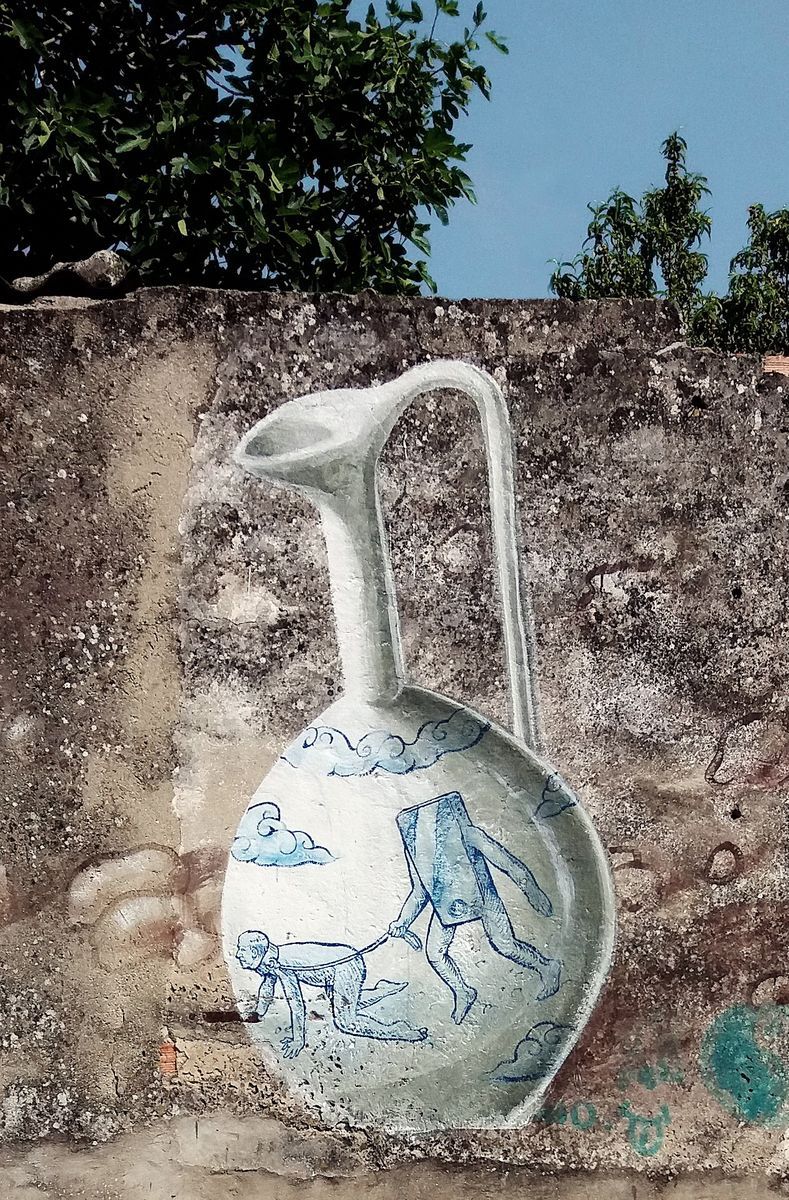 Close-up of a mural depicting a tall white ceramic jug decorated in blue with a figure leading a rabbit-eared animal on a leash, painted on a rough stone wall beneath tree branches.