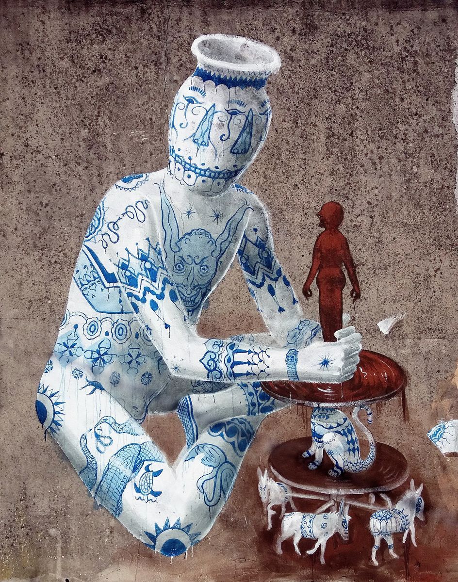 Close-up of a mural showing a seated human figure whose body is entirely covered in blue-and-white ceramic-style patterns and a vase-shaped head, working a potter's wheel that holds a small reddish-brown human figure and miniature goats.