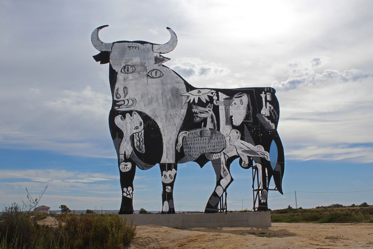 Wide daytime view of a large flat cutout mural styled after Picasso's Guernica bull standing in a dry field under a cloudy sky, its body filled with the painting's horse and human figures rendered in black and white.