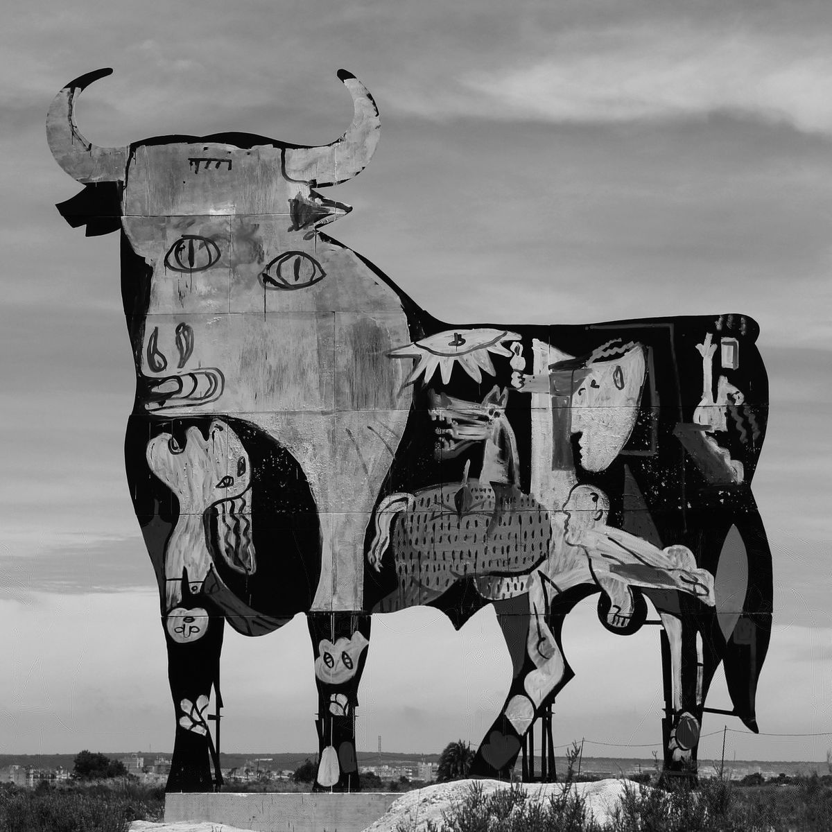 Grayscale photograph of the same Guernica-style bull cutout mural, shown in full against a cloudy sky with distant rooftops visible at its base.