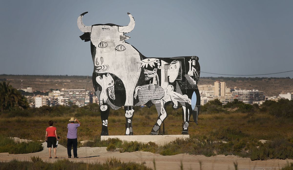 Large flat cutout mural in a scrubland field, styled after Picasso's Guernica bull, painted in black and white with a horse and human figures worked into its body, two visitors viewing it with a city skyline behind.