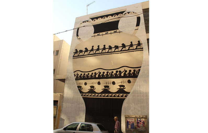 Full building facade mural of a giant black-figure Greek amphora, showing a row of seated figures and its decorative bands, a car and a pedestrian at street level.