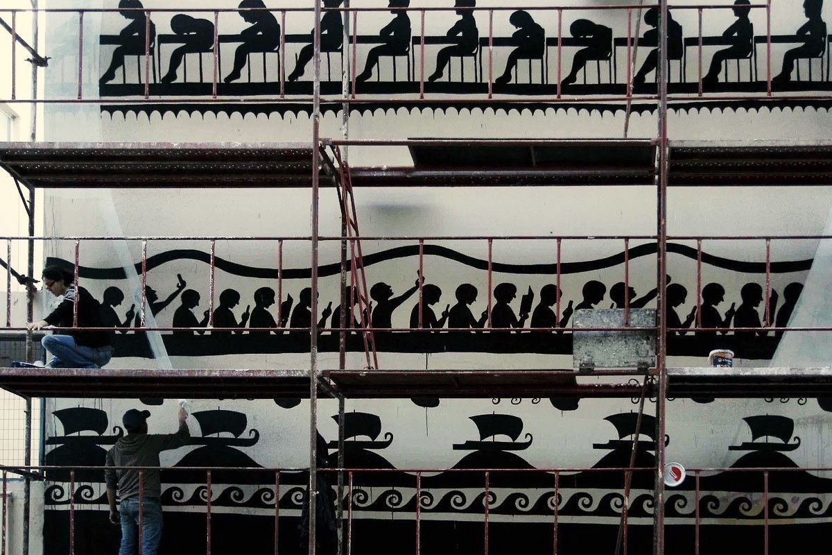 View of the amphora mural through construction scaffolding, showing the row of seated figures, a wavy band of figures raising phones, and a row of ships above a wave pattern, with two painters working on the wall.