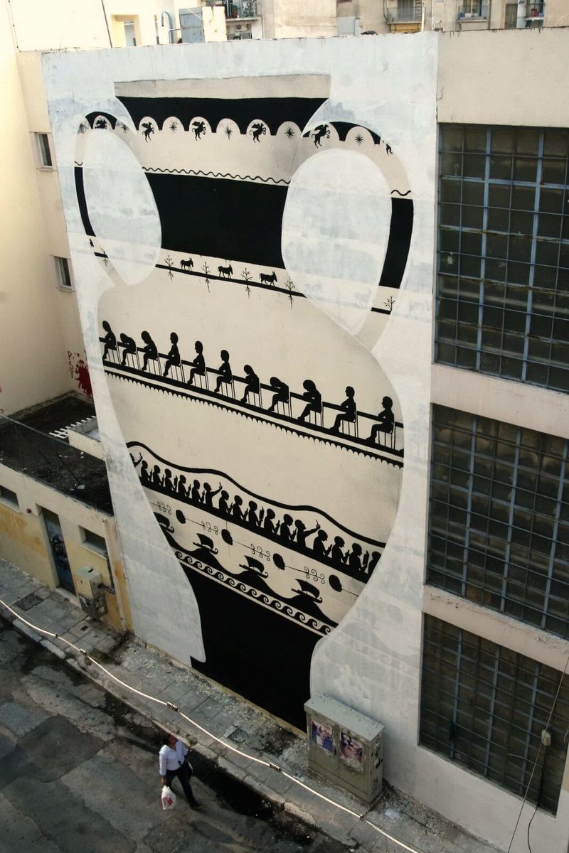 Mural on a building facade depicting a giant Greek amphora with black-figure pottery patterns, including a frieze of seated figures and a row of dancing figures.