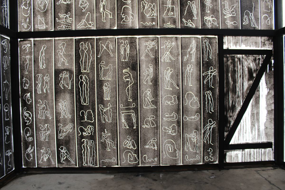 Wall of ink drawings on paper backlit in a glass display case, showing columns of loosely sketched human figures in various poses.