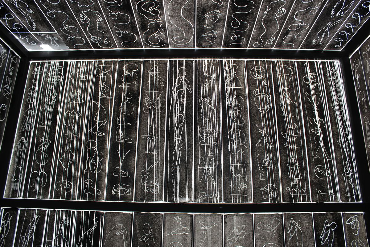 Corner view of a room lined with backlit ink drawings on paper, showing rows of abstract figures, faces and animal-like shapes covering the walls and ceiling.