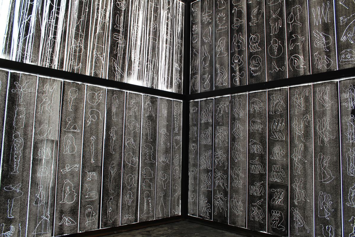 Corner installation of tall dark panels engraved with dozens of small white outline figures and pictograms in a loose grid, illuminated from behind, one upper section streaked with white drips.