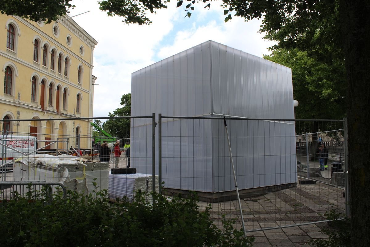 Large white translucent polycarbonate cube structure under construction, enclosed by a metal fence, with workers nearby and a yellow building in the background.