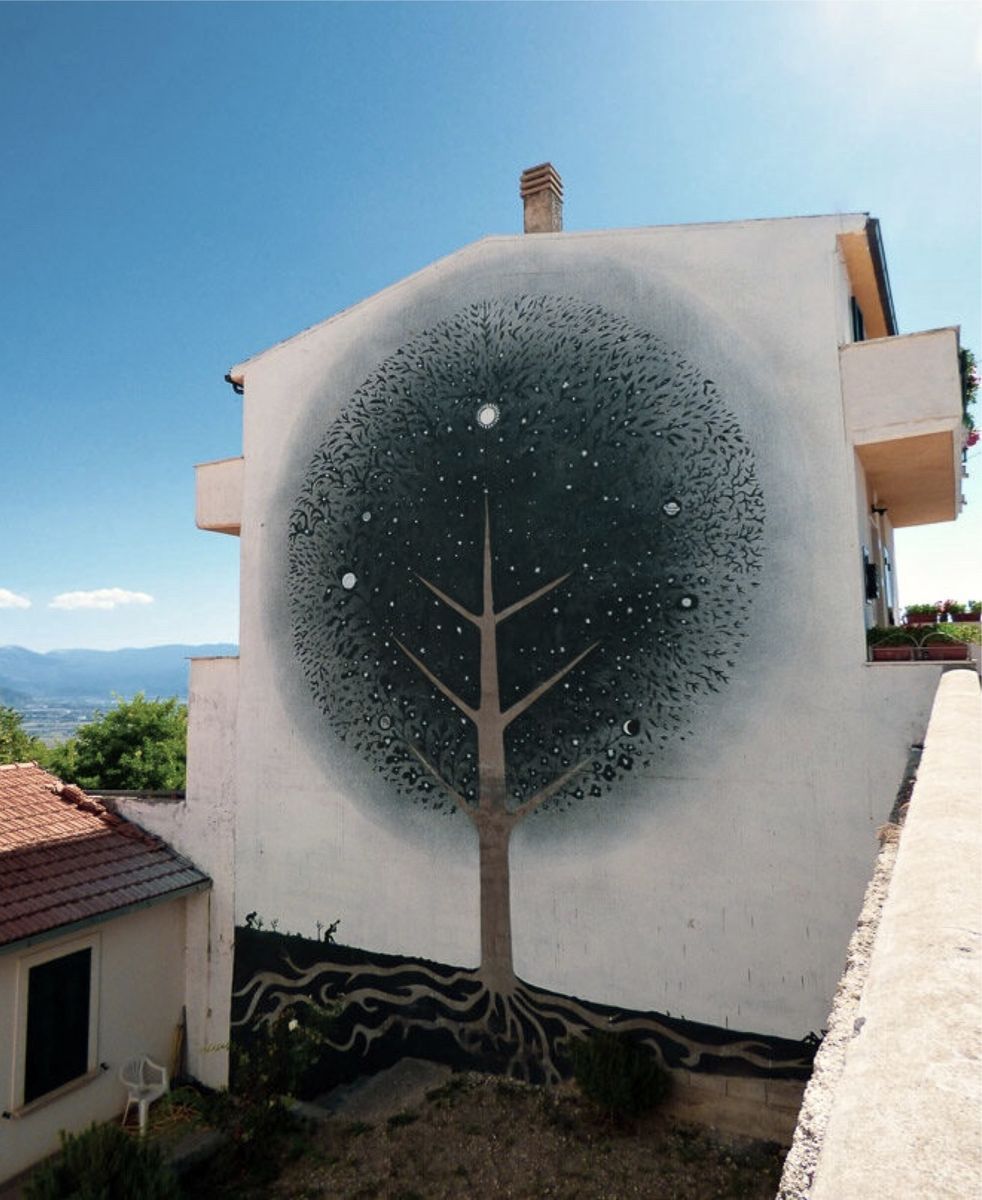 Black silhouette tree with a circular canopy filled with stars, moons, and small flowering shapes, painted on the gable end of a house, its roots spreading into a dark band at the base, mountains visible in the distance.