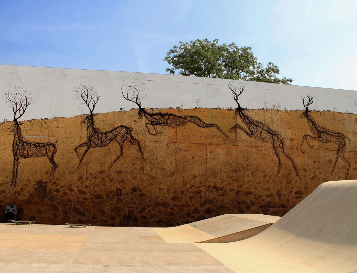 Wide view of five black wireframe deer made of root lines walking across a long wall, white sky above and ochre earth below, a green tree visible behind the wall, two skateboards resting on the ground.