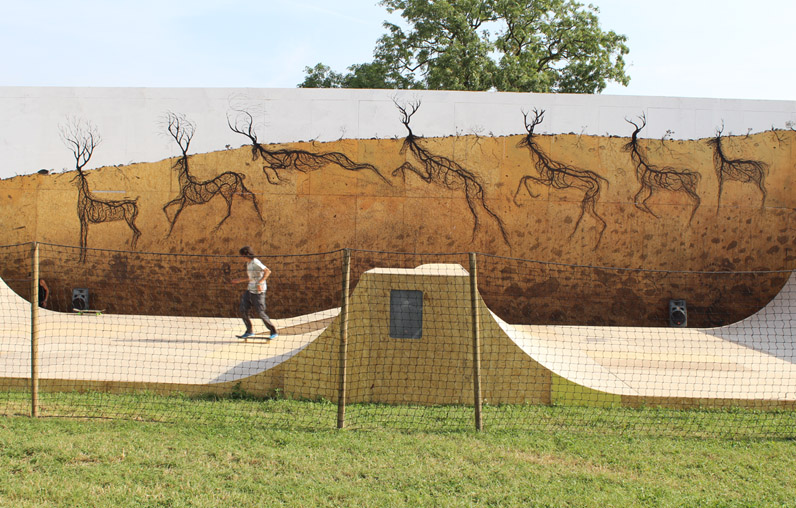 Row of black wireframe deer figures made from tangled root-like lines, running along a raised earthen band on a curved wall above a skate park ramp, with a skateboarder passing below.