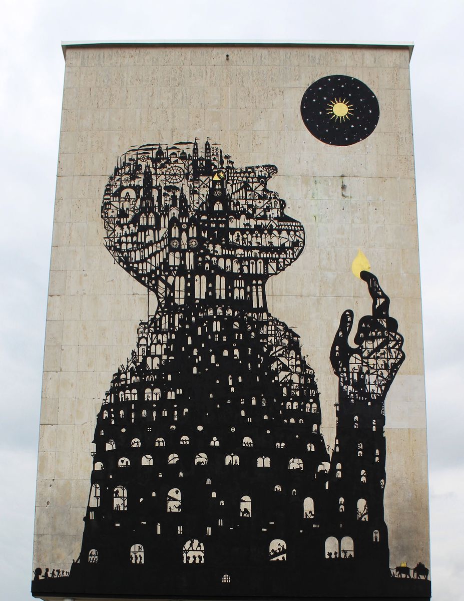 Full view of a tall building facade painted with a black silhouette figure whose head and body are built from an intricate skyline of towers, arches, and staircases, holding up a small yellow flame, beneath a circular black moon with a yellow sun, seen against a cloudy sky.