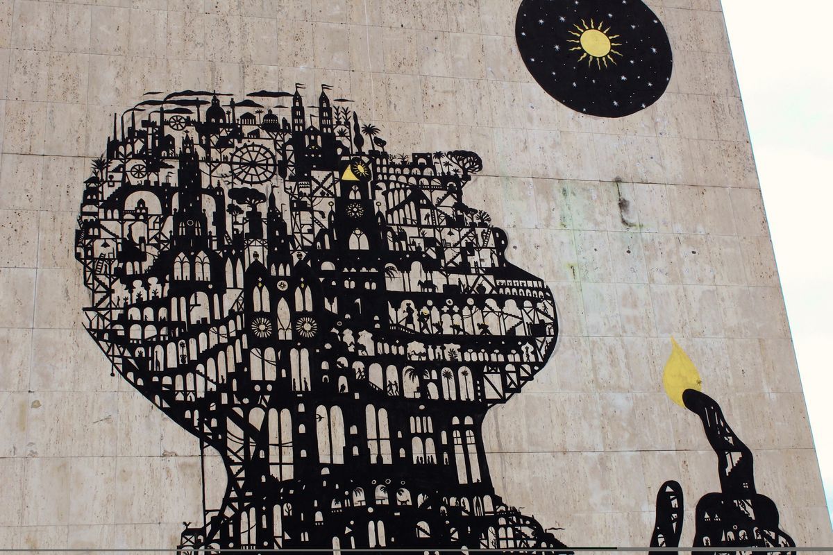 Elevated close view of the building mural showing the detailed cityscape-silhouette head and raised hand with flame, with a residential street, parked cars, and red-roofed houses visible below.