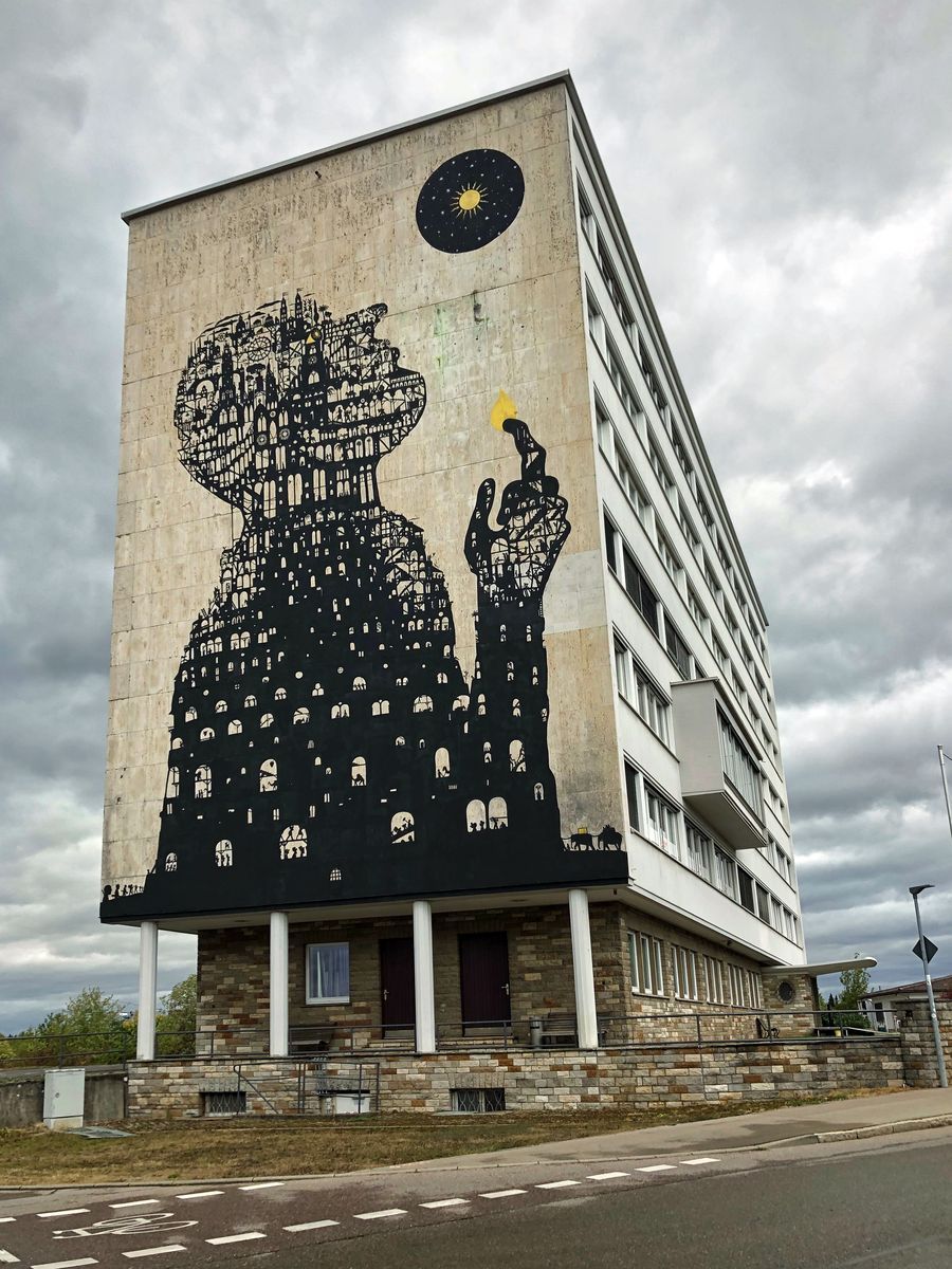 Black silhouette of a robed figure with a head and body built from densely stacked miniature buildings, holding up a small flame, painted on the tall side of an apartment block beneath a circular black moon-and-sun motif.