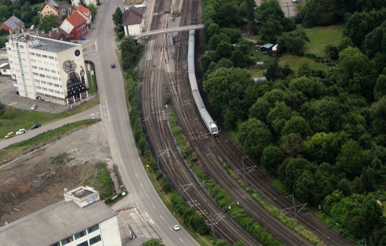 Aerial view of the same building mural showing the black silhouette figure on its facade, set beside curving railway tracks and a passing train, surrounded by trees and a road.