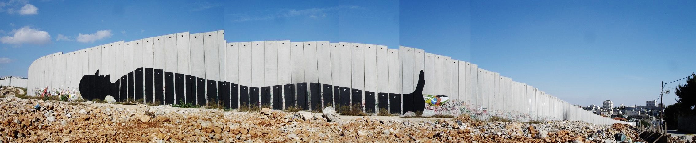 Panoramic view of a long concrete separation wall painted with a black silhouette profile head and an upraised thumb, the shape stretched across dozens of wall panels, rubble-strewn ground in front.