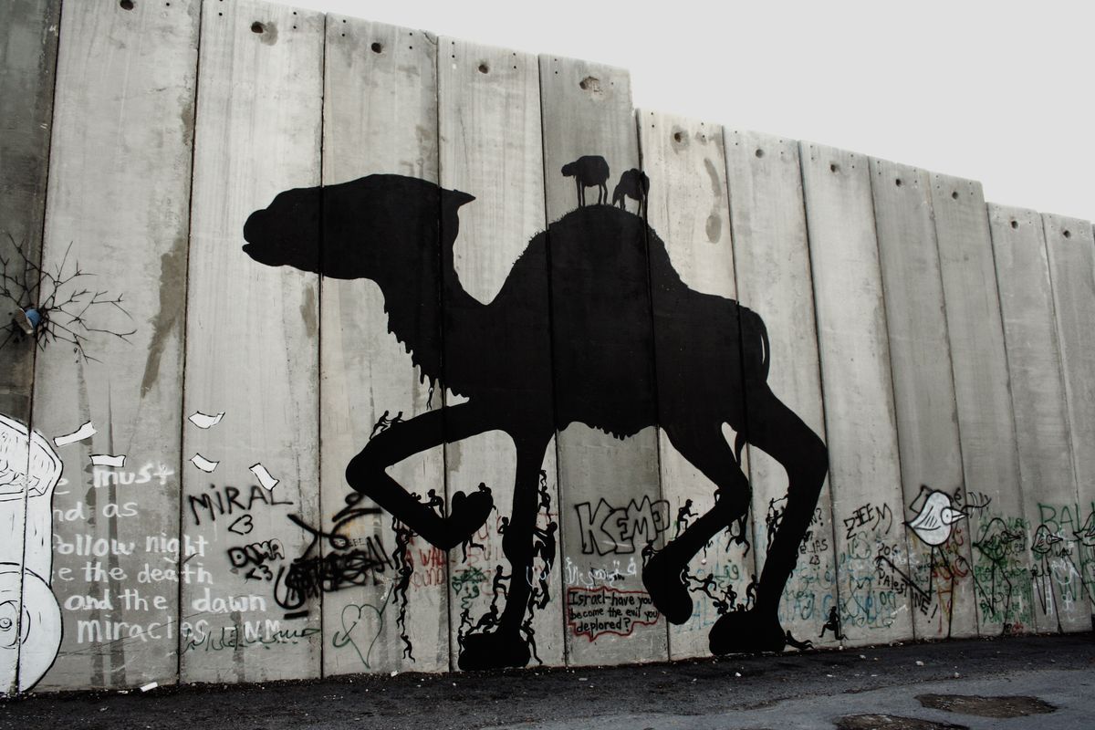 Large black silhouette of a camel painted on a concrete separation wall panel, two small animals standing on its hump, tiny climbing figures and graffiti text scattered across its legs and the surrounding panels.