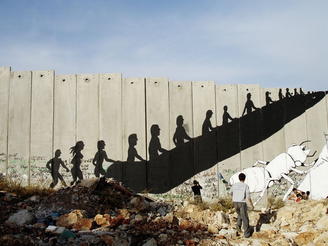 Row of black silhouette figures ascending a diagonal escalator-like line painted across a concrete separation wall, a white livestock animal painted at the base, rubble and children in the foreground.