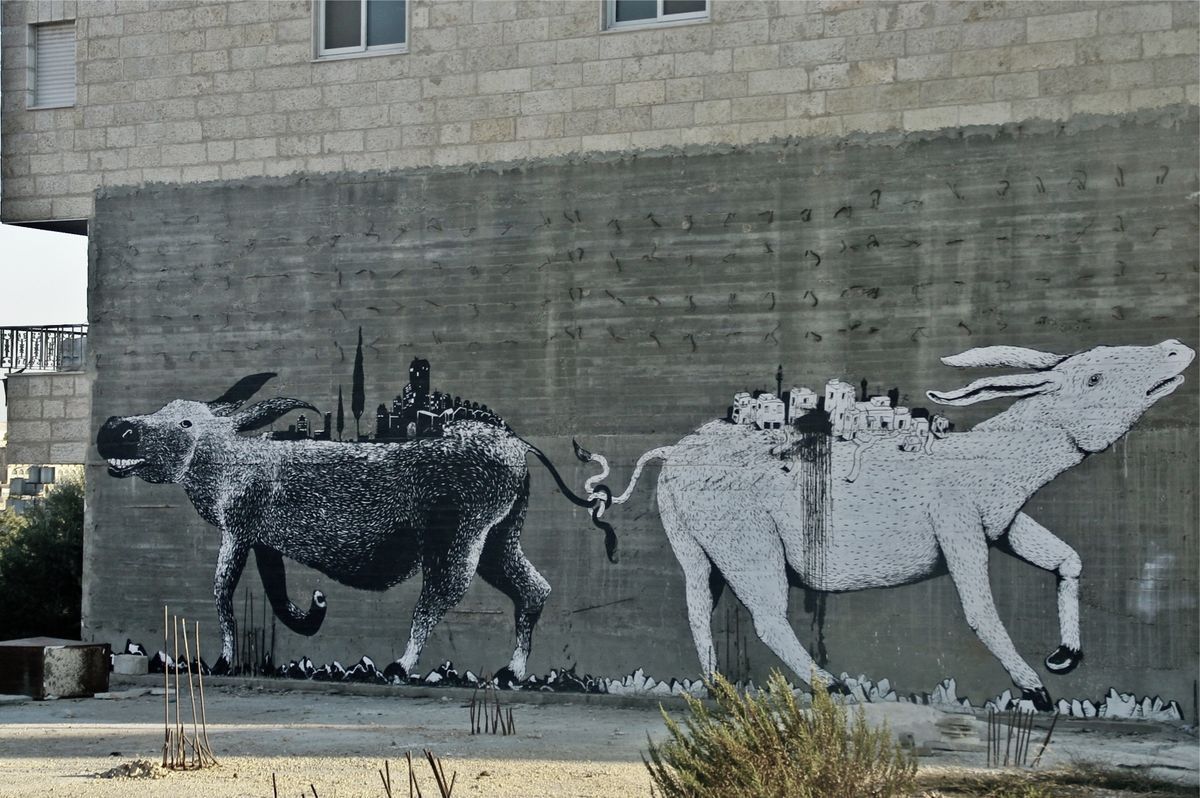 Two black-and-white donkeys facing away from each other on a concrete wall, each carrying a miniature hilltop town on its back, their tails intertwined with a snake between them, birds scattered across the wall.