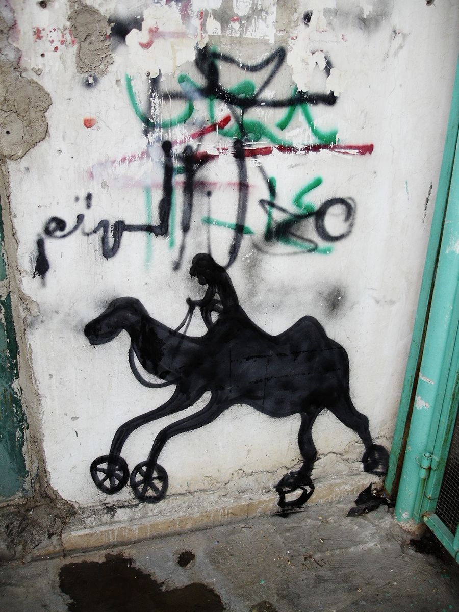 Black silhouette stencil of a camel fitted with small wheels on its front legs, a robed rider holding the reins, spray-painted on a weathered plastered wall beside a teal metal door.