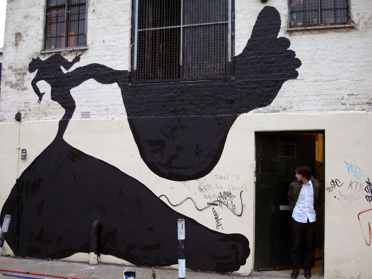 Giant black silhouette of an upturned foot and leg spanning a building facade, a tiny falling female figure at the toes, barred windows above, a man standing in a doorway below.