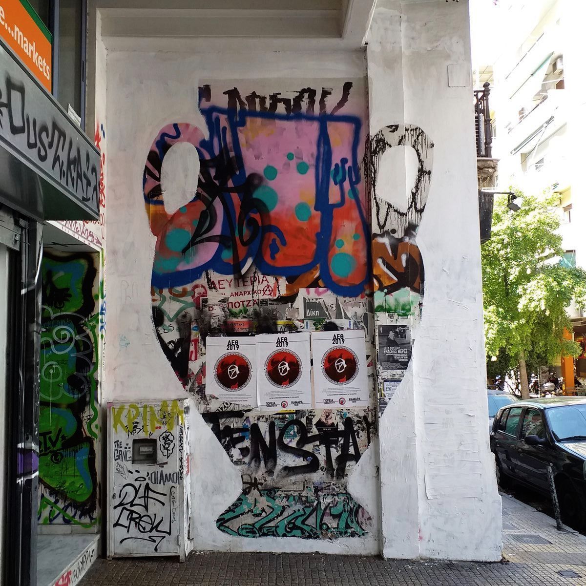 Close-up of a white amphora-shaped outline painted over a wall covered in colorful graffiti tags, stickers, and posters, framed by parked cars and a tree-lined street.