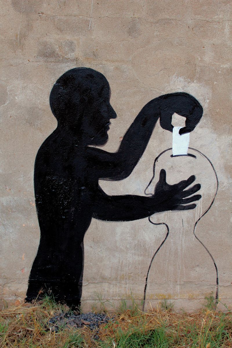 Black silhouette of a man shaping the head of a humanoid figure with his hands, the figure's lower body rendered as a plain outlined amphora shape, painted on a weathered stone wall amid dry grass.