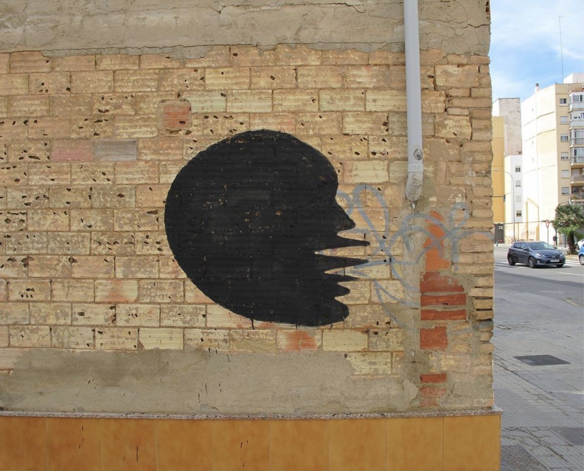 Black rounded silhouette with a jagged, tooth-like edge on one side, painted over a weathered brick wall pocked with small holes, beside a white drainpipe and faint blue graffiti scribbles.