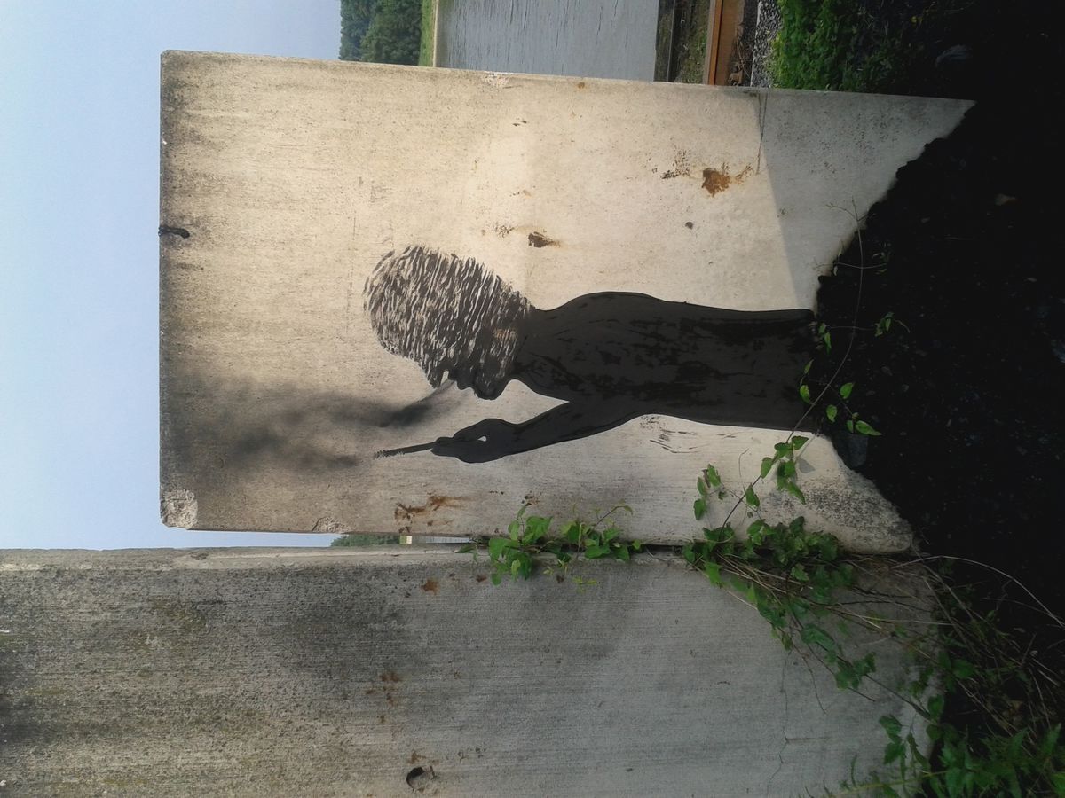 Black silhouette of a seated figure smoking a cigarette, exhaling a trail of smoke, painted on a concrete slab beside a low wall, framed by climbing green vines.