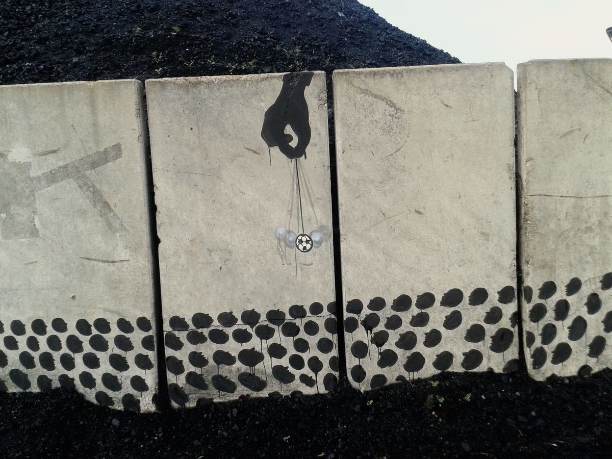 Black silhouette of a hand dangling several small balls on strings, including a black-and-white soccer ball, above a row of concrete panels patterned with dark dripping dots along the bottom edge, a pile of dark gravel behind.