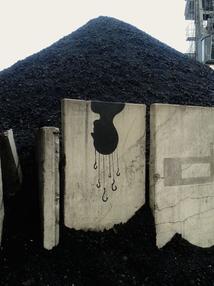 Black silhouette of a head in profile with several fishing hooks dangling from its chin on thin lines, painted on a marble-veined concrete panel, set against a large mound of dark gravel and industrial structures.