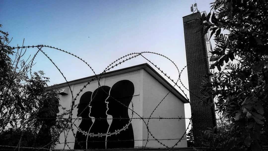 Four black silhouette profile heads in a row on a white building facade, connected mouth to mouth by strands of barbed wire, photographed through overhanging branches and coils of barbed wire in the foreground.