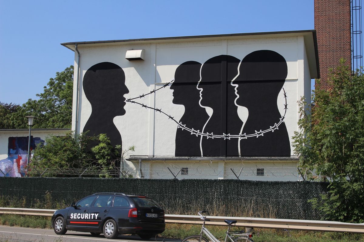 Wide view of four black silhouette profile heads painted on a white building, linked together by a strand of barbed wire running from mouth to mouth, with a security car and bicycle parked on the street below.