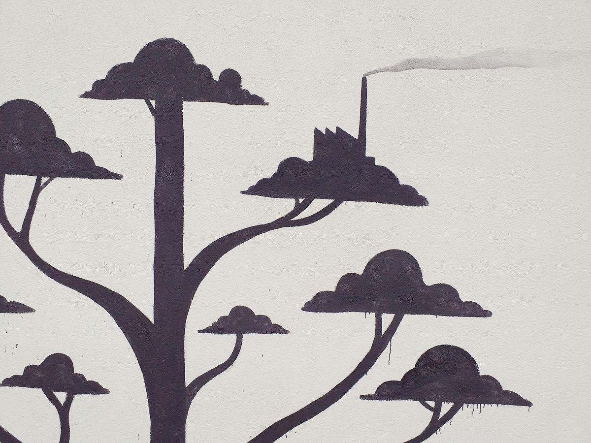 Detail of black silhouette trees with cloud-shaped canopies, one canopy shaped like a small factory with a chimney releasing a trail of smoke, painted on a plain wall.
