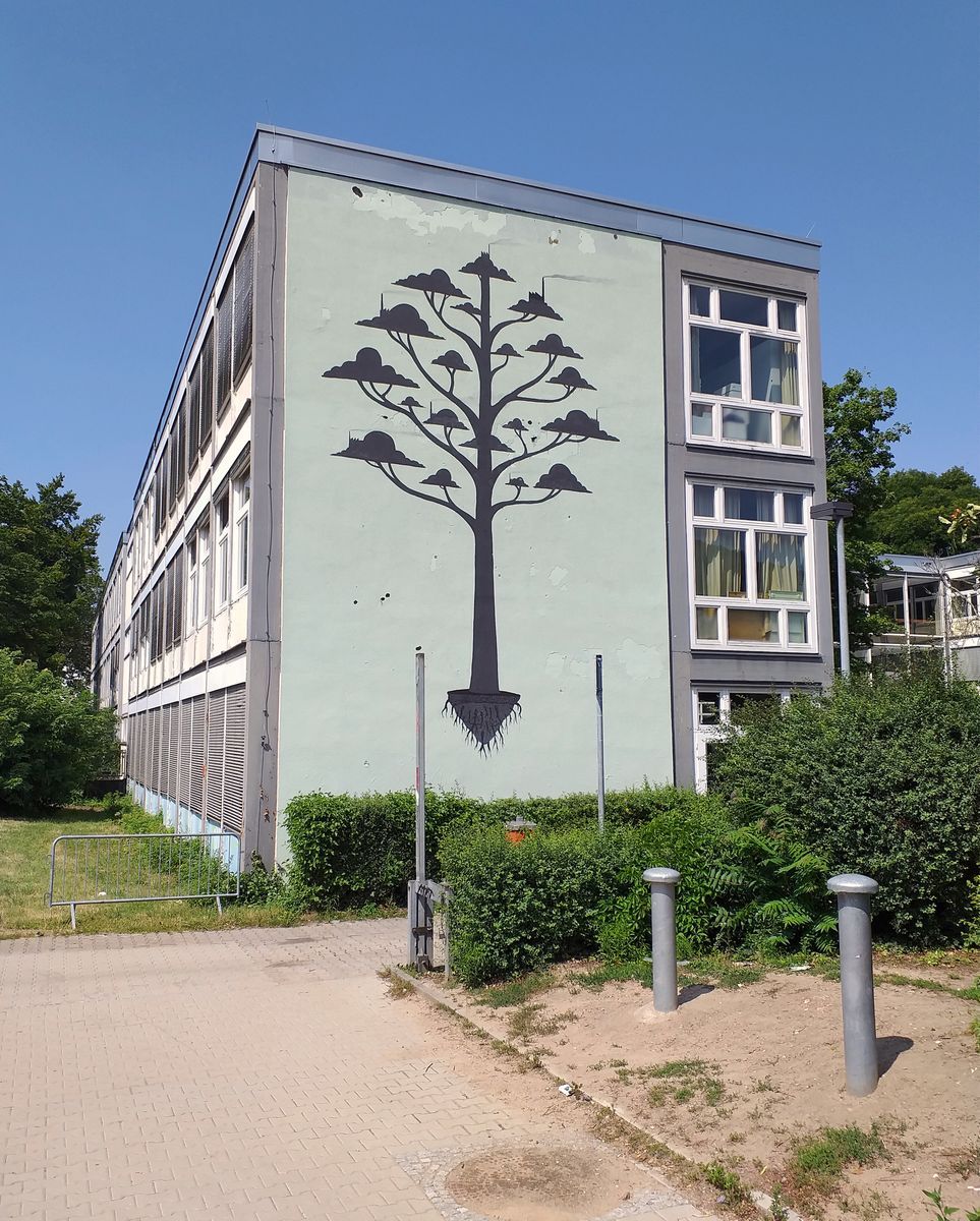 Full black silhouette of a tree with multiple cloud-shaped canopies branching from a single trunk and exposed roots below, painted on the corner of a school building beside a row of windows.